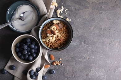 Photo of Tasty granola in bowl, blueberries, yogurt and spoon on gray textured table, flat lay. Space for text