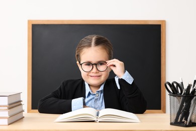 Happy little school child sitting at desk with books near chalkboard in classroom