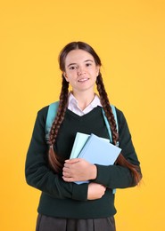 Teenage girl in school uniform with books and backpack on yellow background