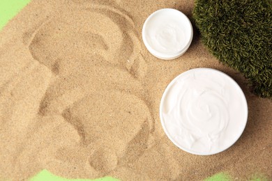 Jars with cream and moss on sand against green background, flat lay with space for text. Cosmetic product