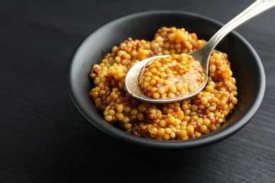 Photo of Whole grain mustard in bowl and spoon on black wooden table, closeup