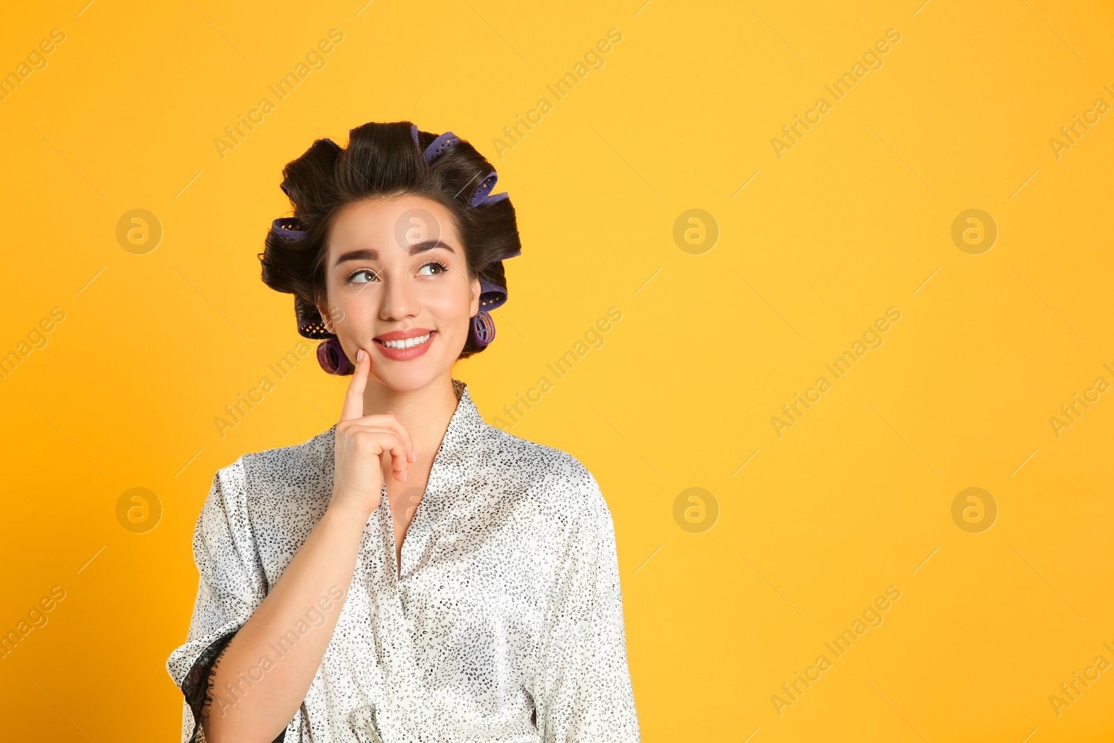 Photo of Happy young woman in silk bathrobe with hair curlers on orange background, space for text