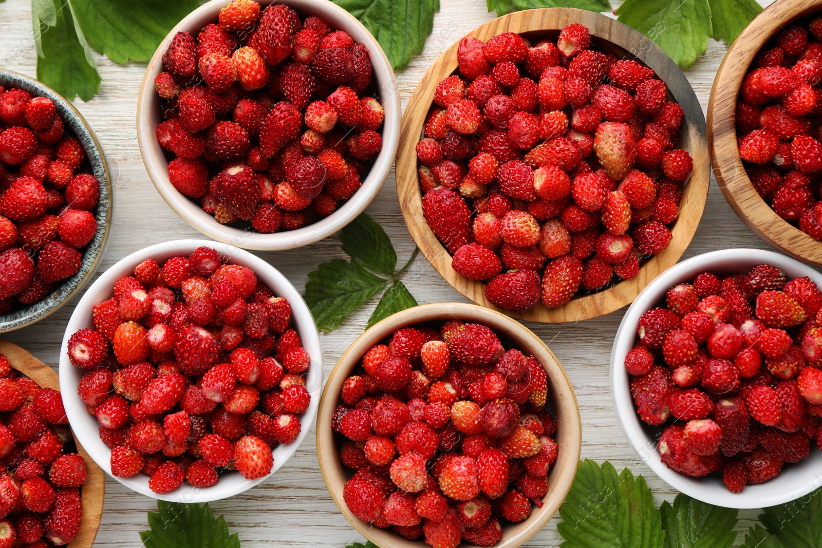 Photo of Fresh wild strawberries in bowls and leaves on white wooden table, flat lay