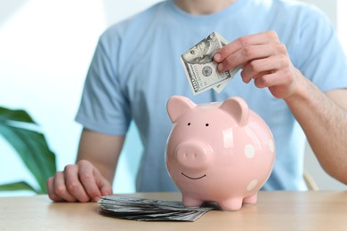 Photo of Financial savings. Man putting dollar banknote into piggy bank at wooden table, closeup