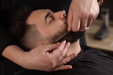 Professional barber shaving client's beard with blade in barbershop, closeup