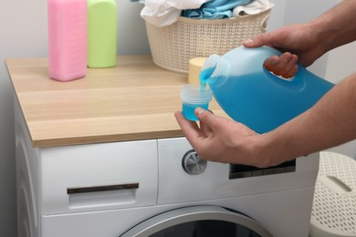 Photo of Man pouring fabric softener from bottle into cap near washing machine indoors, closeup
