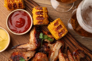 Photo of Delicious baked chicken wings, grilled corn, sauces and glasses of beer on wooden table, flat lay