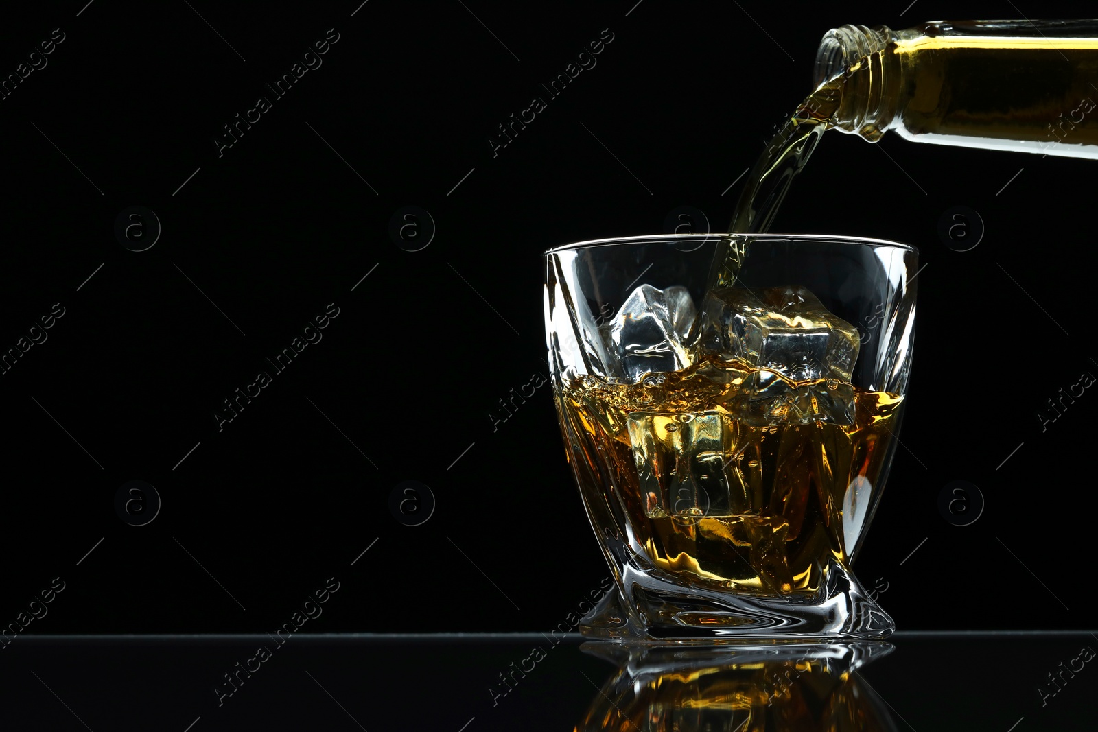 Photo of Pouring whiskey from bottle into glass with ice cubes at table against black background, space for text