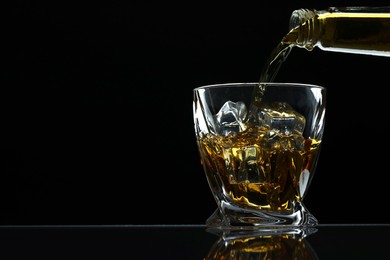 Pouring whiskey from bottle into glass with ice cubes at table against black background, space for text