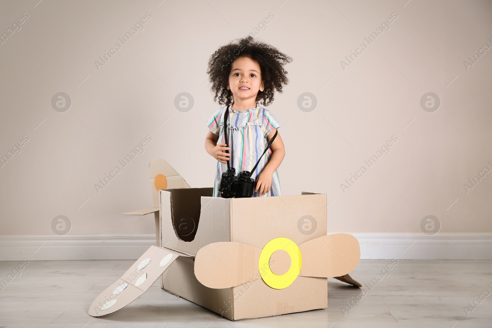 Photo of Cute African American child playing with cardboard plane and binoculars near beige wall
