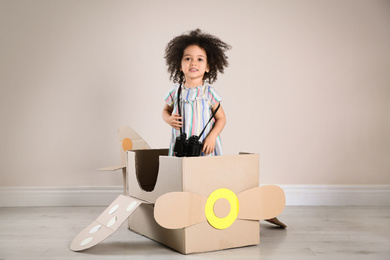 Photo of Cute African American child playing with cardboard plane and binoculars near beige wall