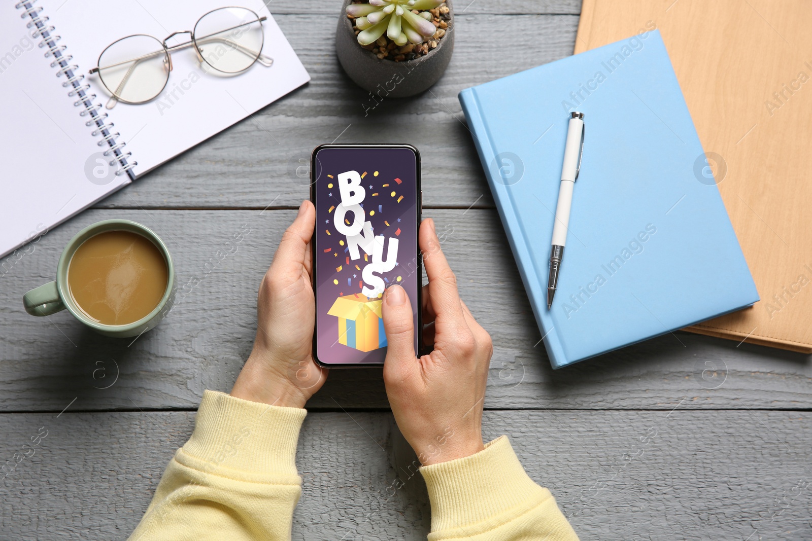 Image of Bonus gaining. Woman holding smartphone at grey wooden table, top view. Illustration of open gift box, word and confetti on device screen