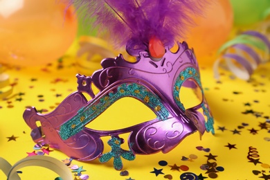 Photo of Beautiful purple carnival mask and shiny confetti on yellow background, closeup