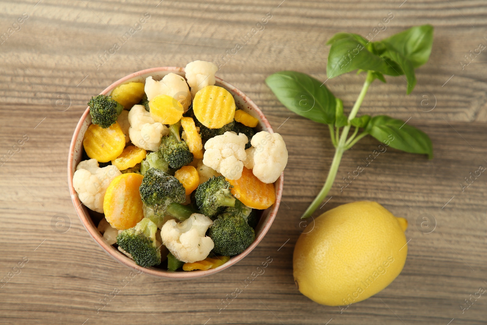 Photo of Frozen vegetables in bowl, basil and lemon on wooden table, flat lay