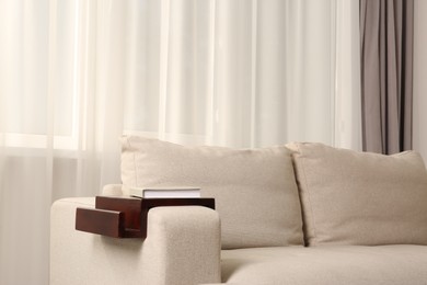 Photo of Wooden armrest table with book on sofa in room. Interior element