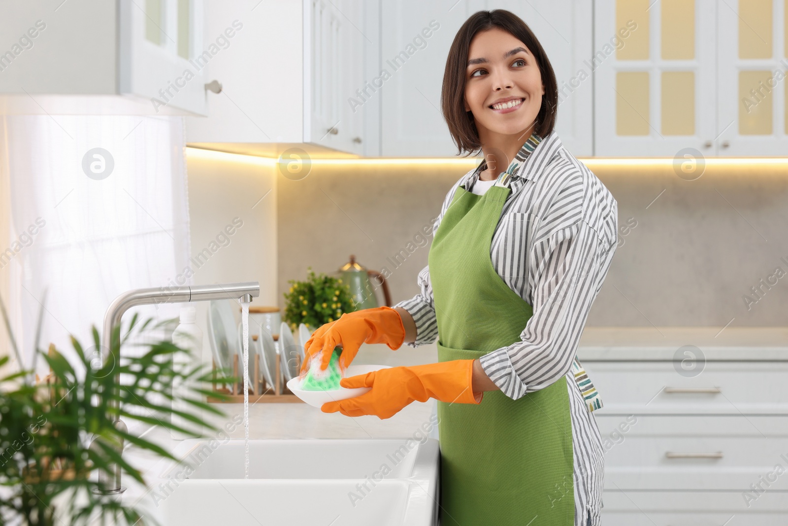 Photo of Happy young woman washing plate above sink in modern kitchen