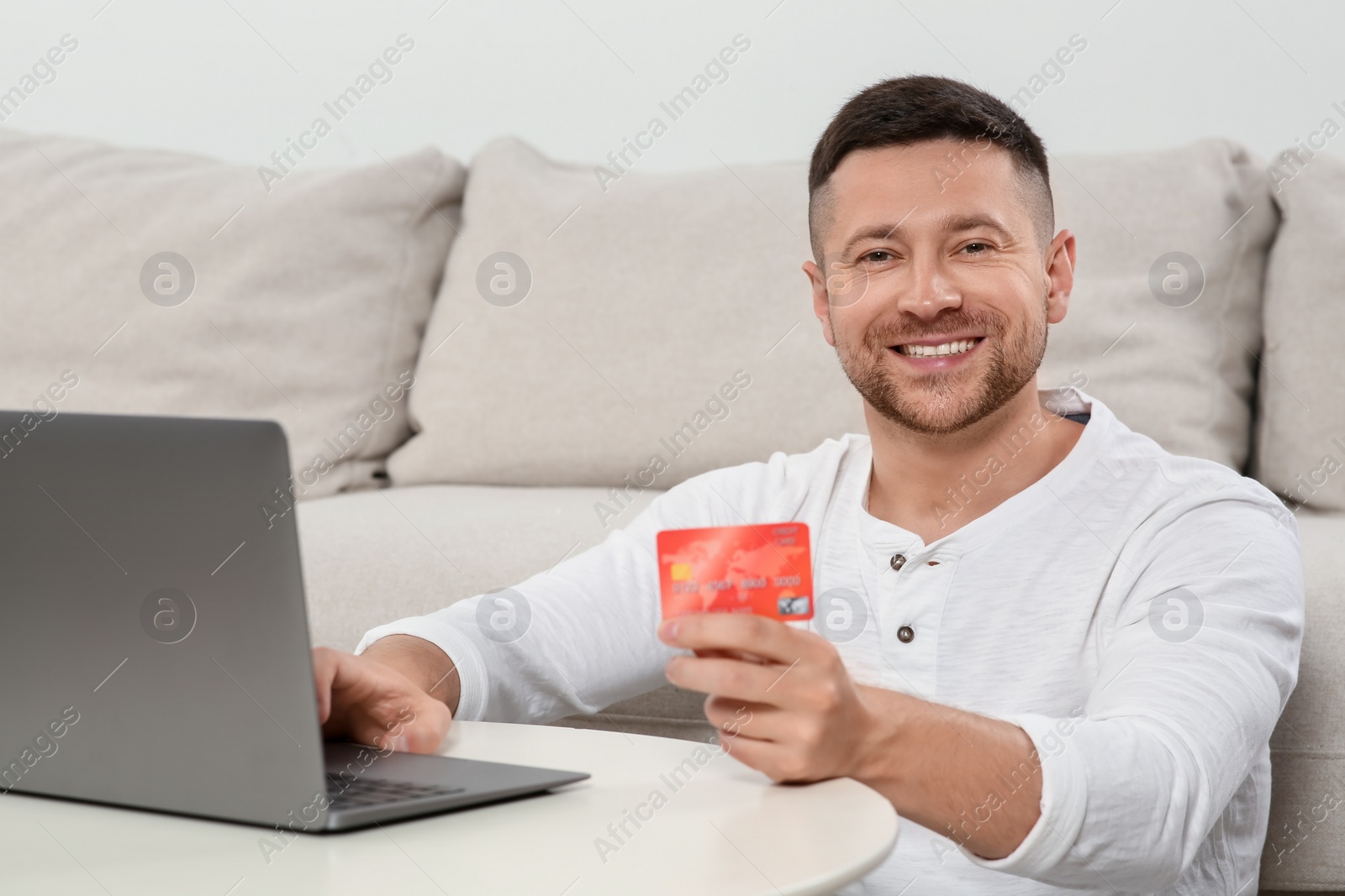 Photo of Happy man with credit card using laptop for online shopping at white table indoors