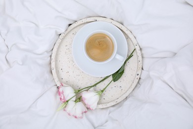 Tray with cup of coffee and flowers on white bed, top view