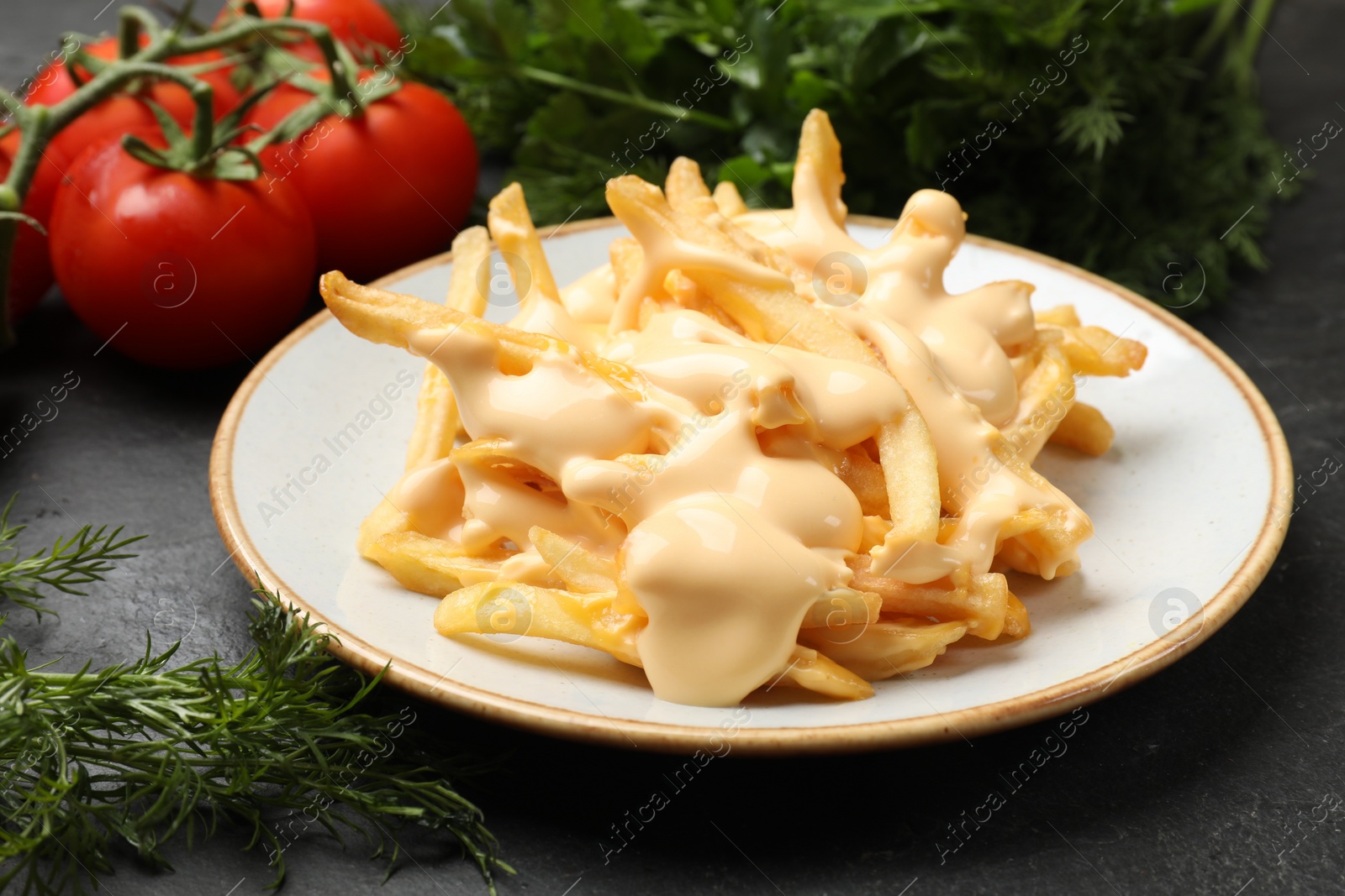 Photo of Delicious French fries with cheese sauce, tomatoes, dill and parsley on black table, closeup