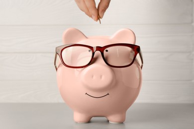 Woman putting coin into piggy bank with glasses at light grey table, closeup