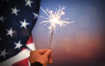 Image of 4th of July - Independence Day of USA. Woman holding burning sparkler near American flag, closeup