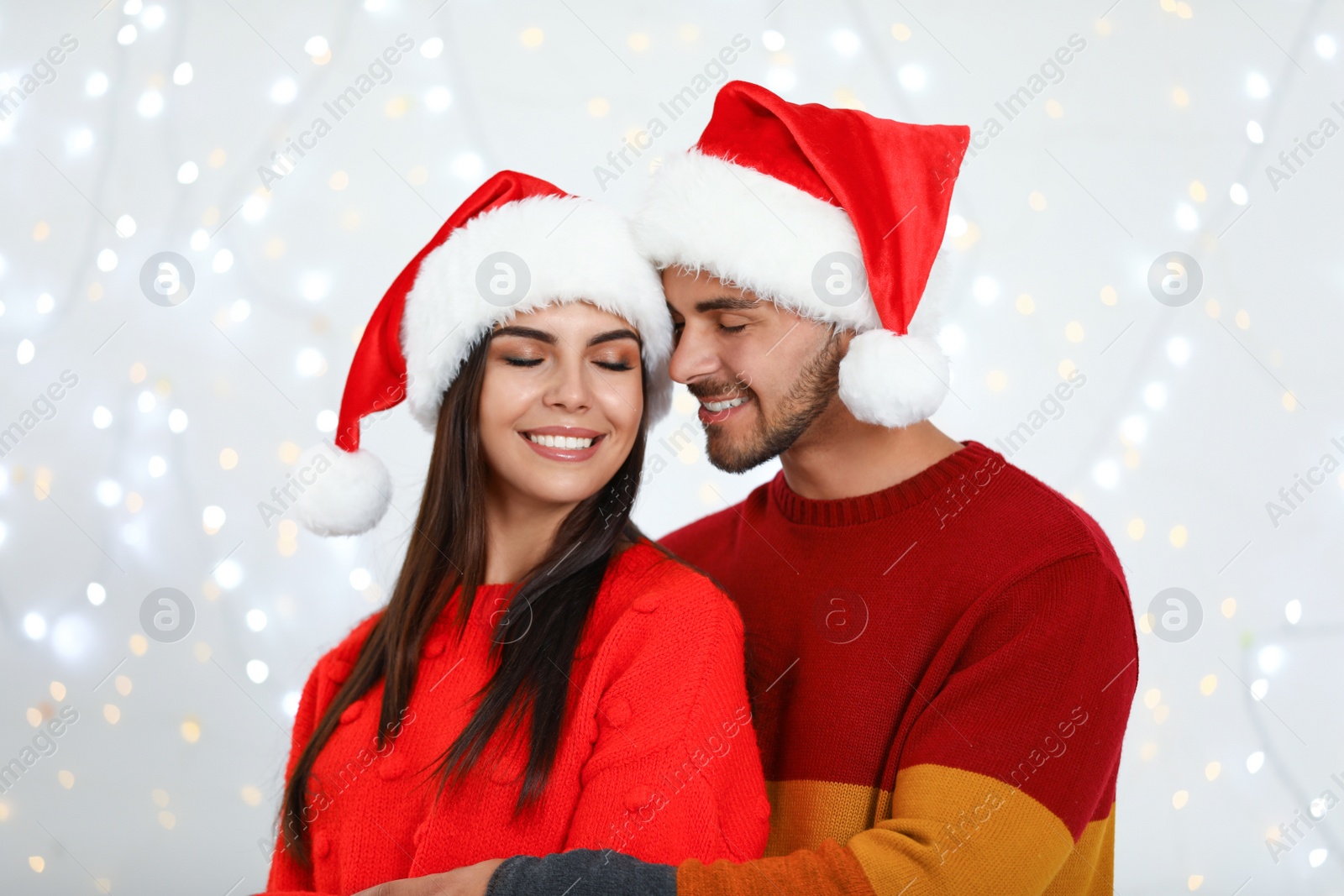 Photo of Lovely young couple in Santa hats against blurred festive lights. Christmas celebration