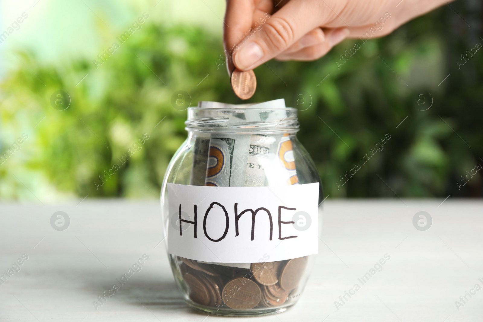 Photo of Woman putting coin into jar with tag HOME on white wooden table, closeup