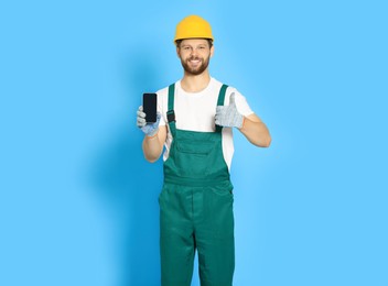 Professional repairman in uniform showing smartphone and thumbs up on light blue background