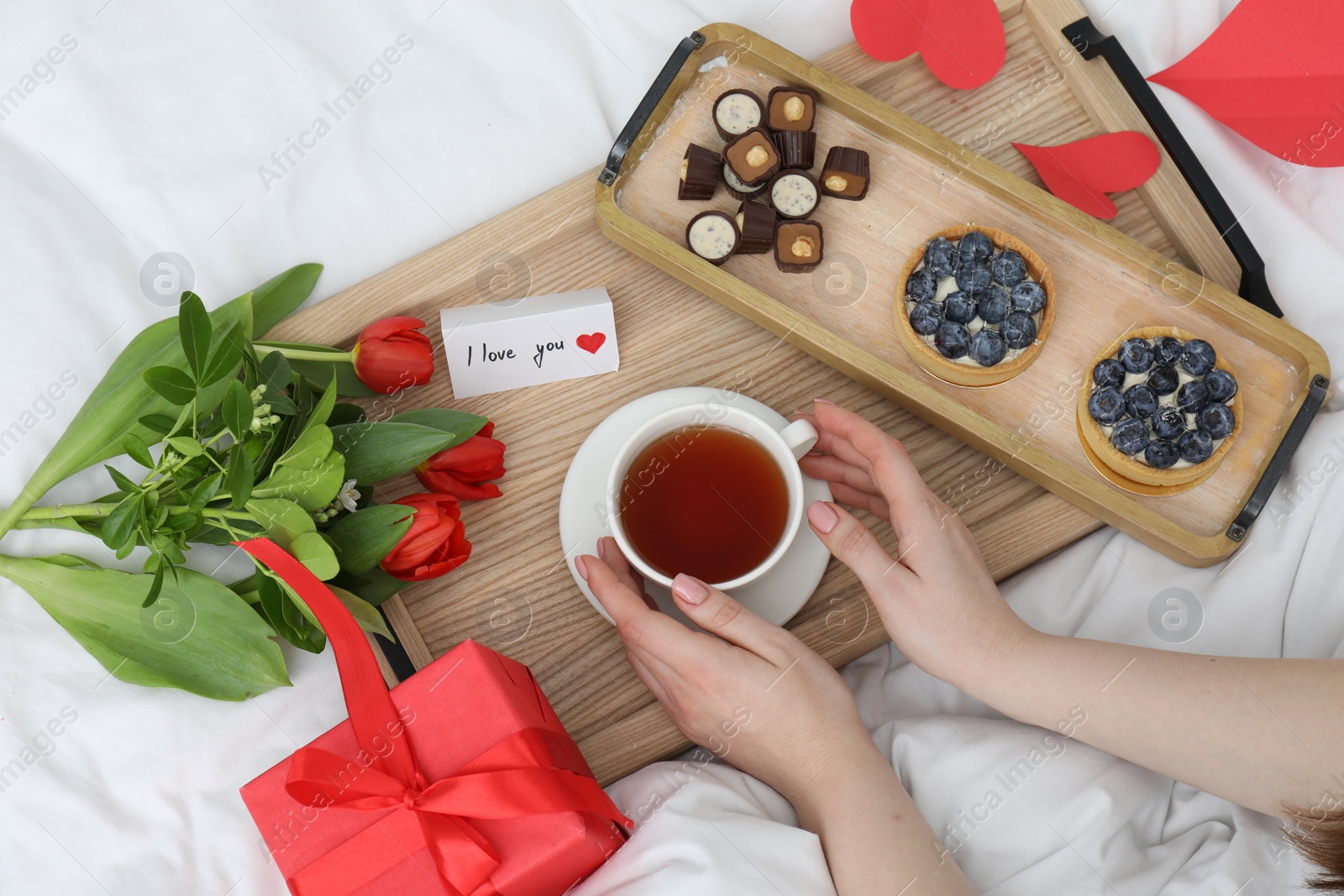 Photo of Tasty breakfast served in bed. Woman with tea, desserts, gift box, flowers and I Love You card at home, top view