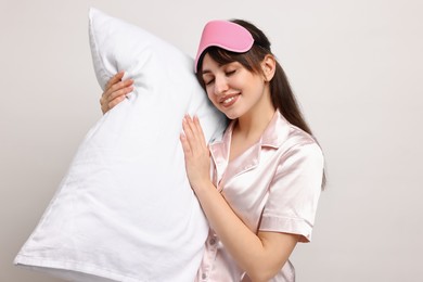 Happy woman in pyjama and sleep mask holding pillow on light grey background
