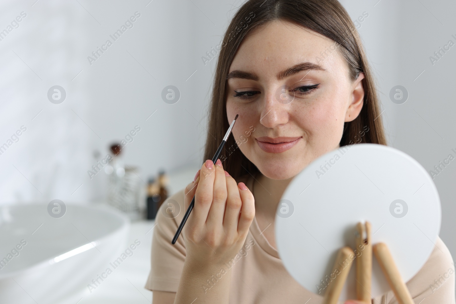 Photo of Beautiful woman drawing freckles with brush in front of little mirror indoors. Space for text