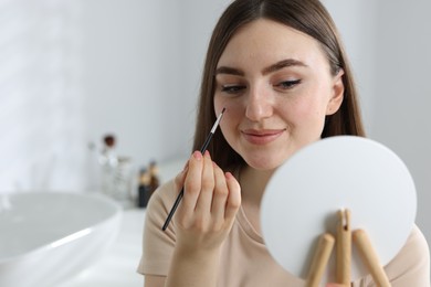 Beautiful woman drawing freckles with brush in front of little mirror indoors. Space for text