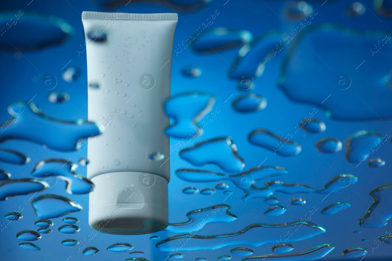 Photo of Moisturizing cream in tube on glass with water drops against blue background, low angle view. Space for text