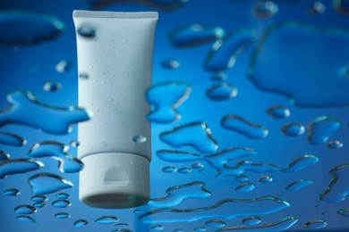 Photo of Moisturizing cream in tube on glass with water drops against blue background, low angle view. Space for text