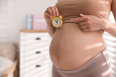 Photo of Young pregnant woman pointing at alarm clock near her belly indoors, closeup. Time to give birth