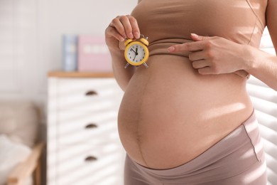 Young pregnant woman pointing at alarm clock near her belly indoors, closeup. Time to give birth