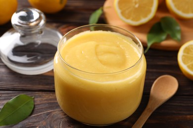 Photo of Delicious lemon curd in glass jar, fresh citrus fruit and spoon on wooden table, closeup