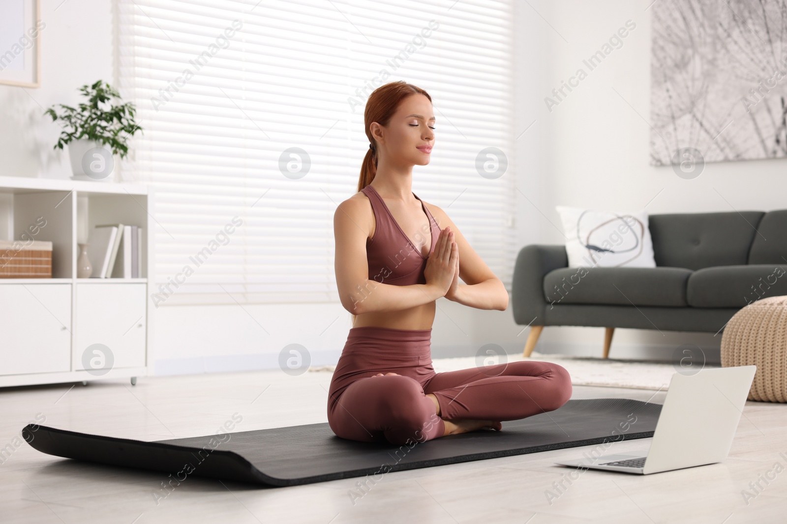 Photo of Beautiful young woman practicing Padmasana with laptop on yoga mat at home. Lotus pose