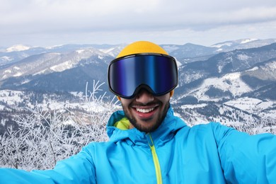 Image of Smiling young man in ski goggles taking selfie in snowy mountains