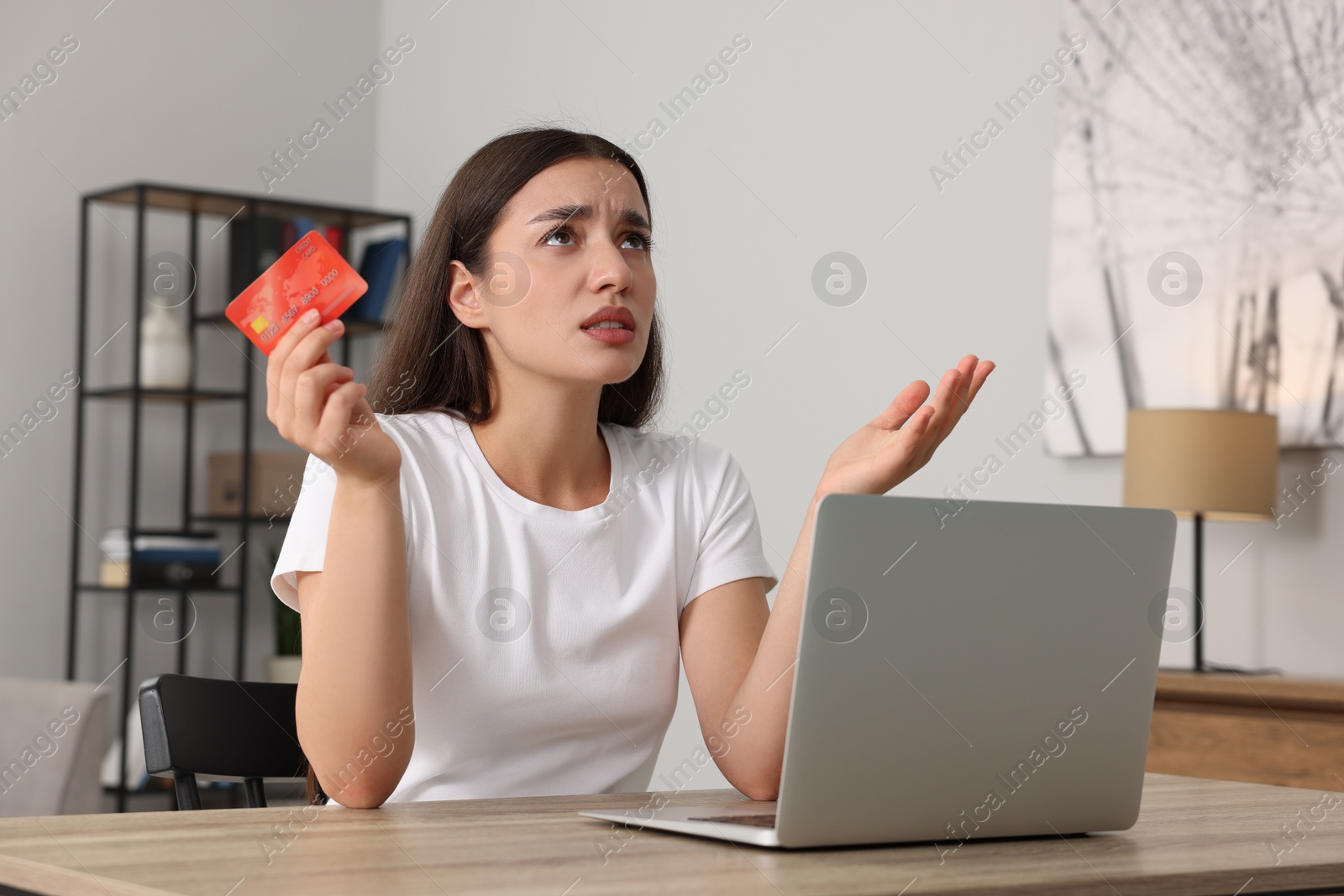 Photo of Confused woman with credit card and laptop at table indoors. Be careful - fraud