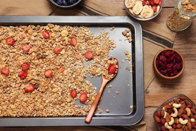 Photo of Making granola. Baking tray with mixture of oat flakes, other ingredients and spoon on wooden table, flat lay