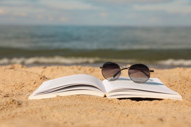 Photo of Open book and sunglasses on sandy beach near sea