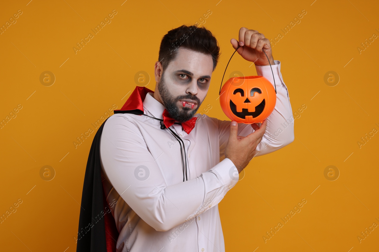 Photo of Man in scary vampire costume with fangs and pumpkin bucket on orange background. Halloween celebration