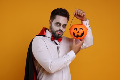 Man in scary vampire costume with fangs and pumpkin bucket on orange background. Halloween celebration