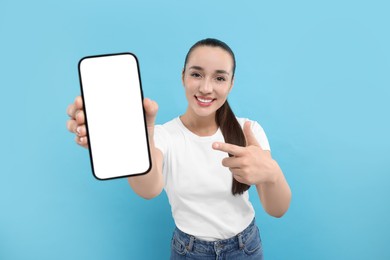 Photo of Young woman showing smartphone in hand and pointing at it on light blue background