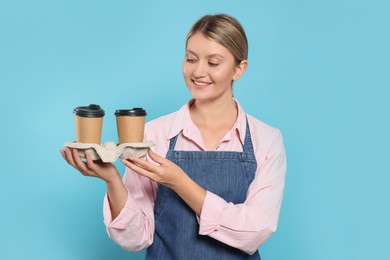 Photo of Beautiful young woman in denim apron with cups of coffee on light blue background