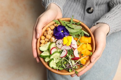 Photo of Woman holding delicious vegan bowl with cucumbers, chickpeas and violet flowers on beige background, closeup