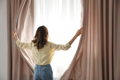 Woman opening window curtains at home in morning, back view