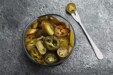 Photo of Glass bowl and fork with slices of pickled green jalapeno peppers on grey table, flat lay