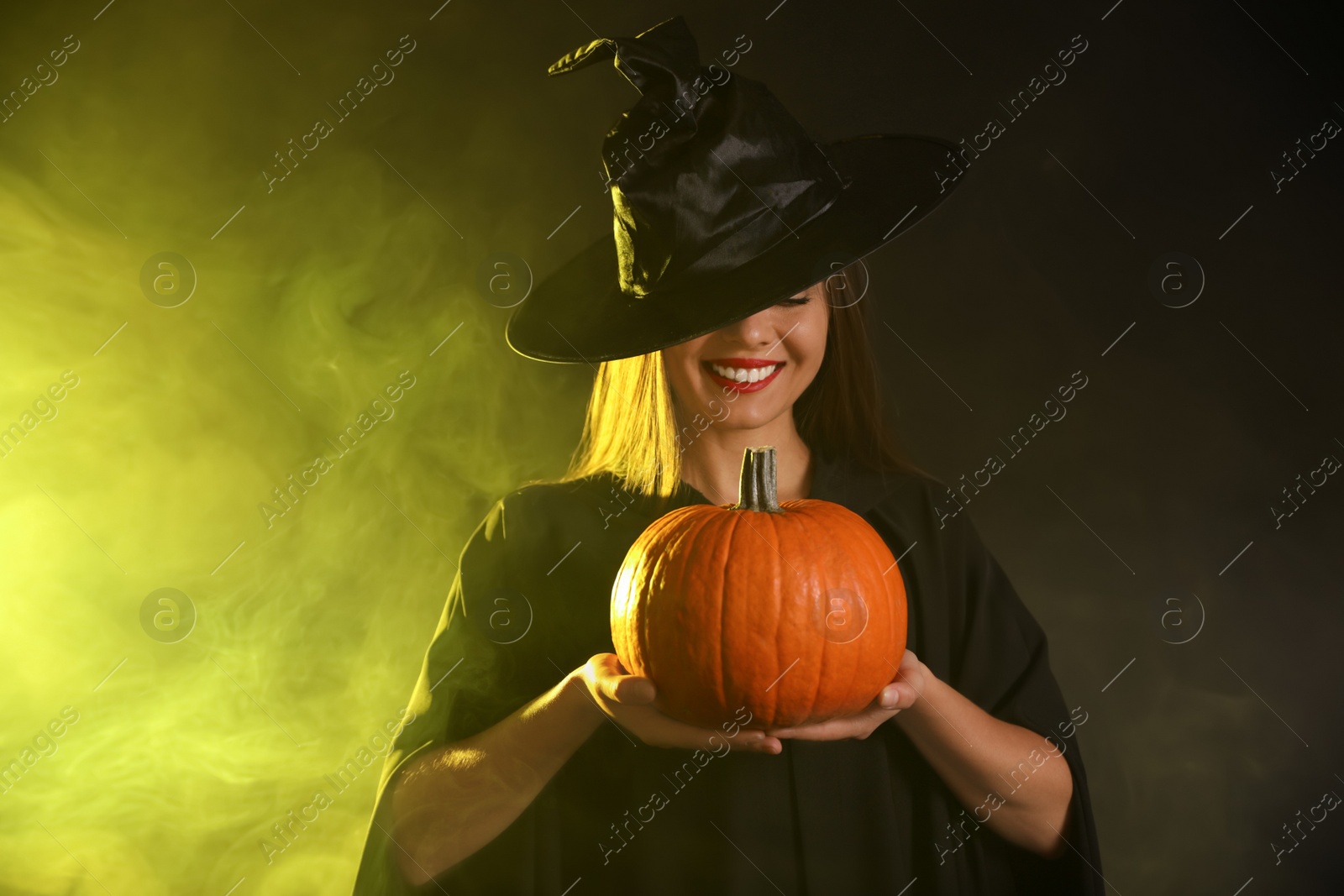 Photo of Young woman wearing witch costume with pumpkin in smoke cloud on dark background. Halloween party